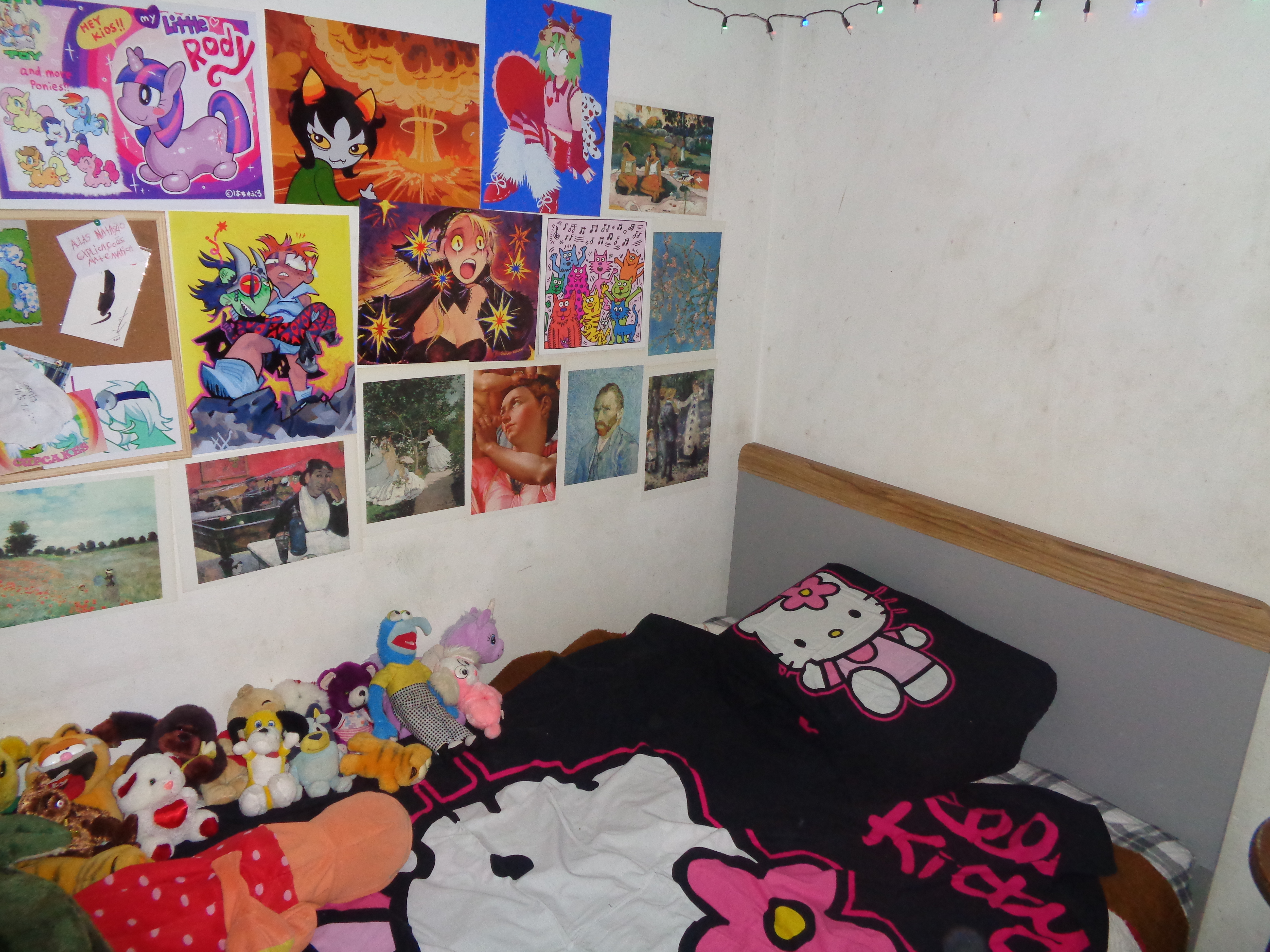 An image of the corner of a bedroom. the bed has hello kitty sheets and is covered in various plushies. above the bed are posters and prints from various artists and medias, such as homestuck, my little pony, dungeon meshi, and more. There are hung up christmas lights just visible in the frame.