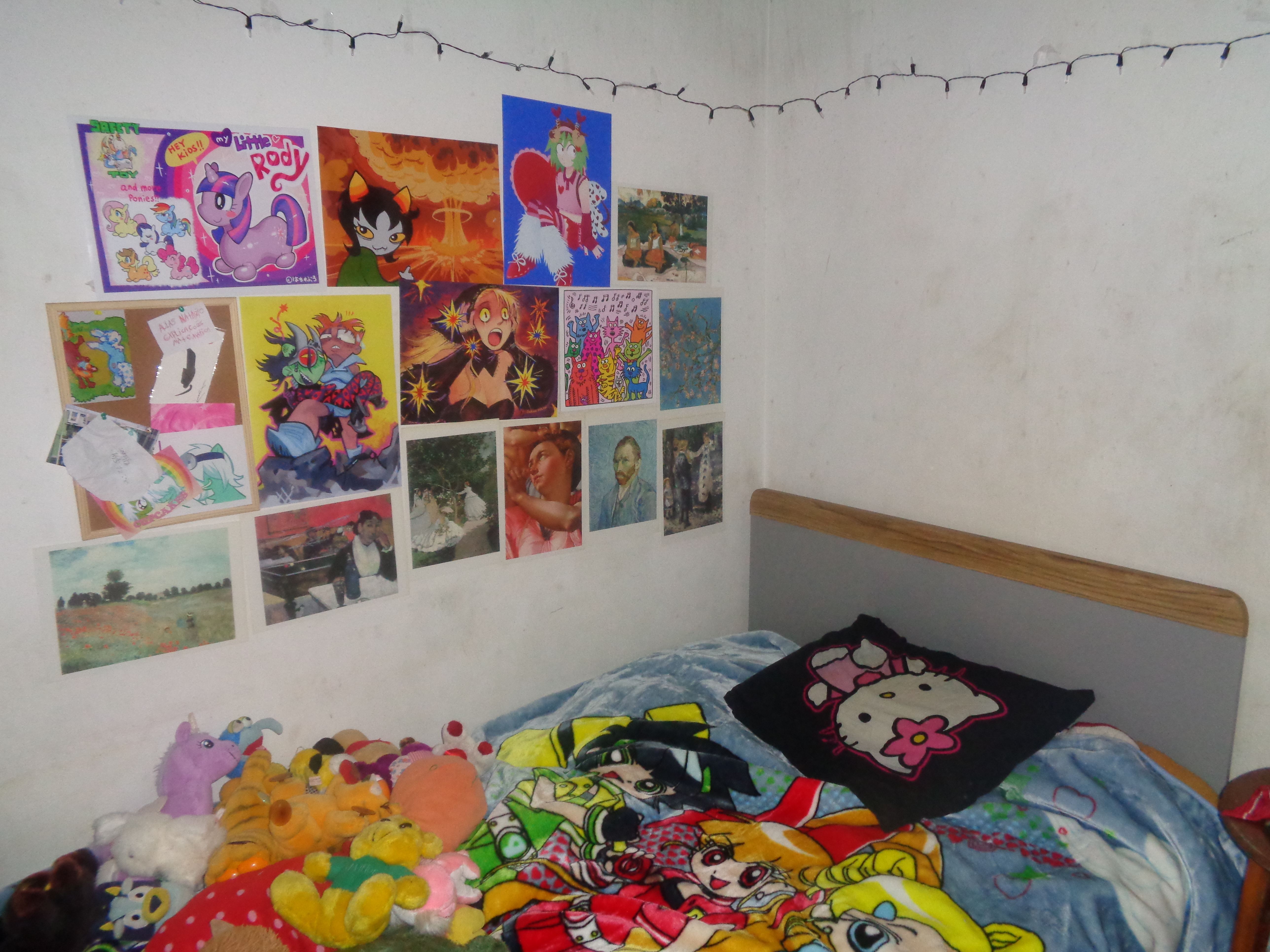 An image of the corner of a bedroom. the bed has powerpuff girls Z sheets and is covered in various plushies. above the bed are posters and prints from various artists and medias, such as homestuck, my little pony, dungeon meshi, and more. There are hung up christmas lights above it all.